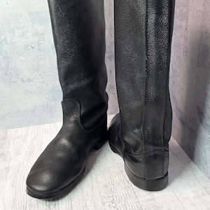 Puede incluir: Un par de botas de cuero negro. Las botas son altas, llegando a la mitad de la pantorrilla, con punta redonda y tac&oacute;n bajo. El cuero tiene un aspecto texturizado. Las botas est&aacute;n sobre una superficie de madera clara.
