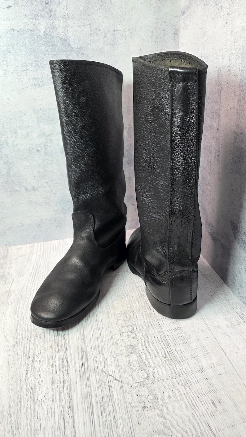 Puede incluir: Un par de botas de cuero negro. Las botas son altas, llegando a la mitad de la pantorrilla, con punta redonda y tac&oacute;n bajo. El cuero tiene un aspecto texturizado. Las botas est&aacute;n sobre una superficie de madera clara.