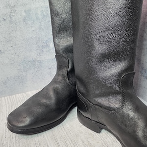 Puede incluir: Un par de botas de montar de cuero negro. Las botas tienen una ca&ntilde;a alta, una puntera redondeada y un tac&oacute;n bajo. El cuero tiene un aspecto texturizado. Las botas est&aacute;n sobre una superficie de madera clara.