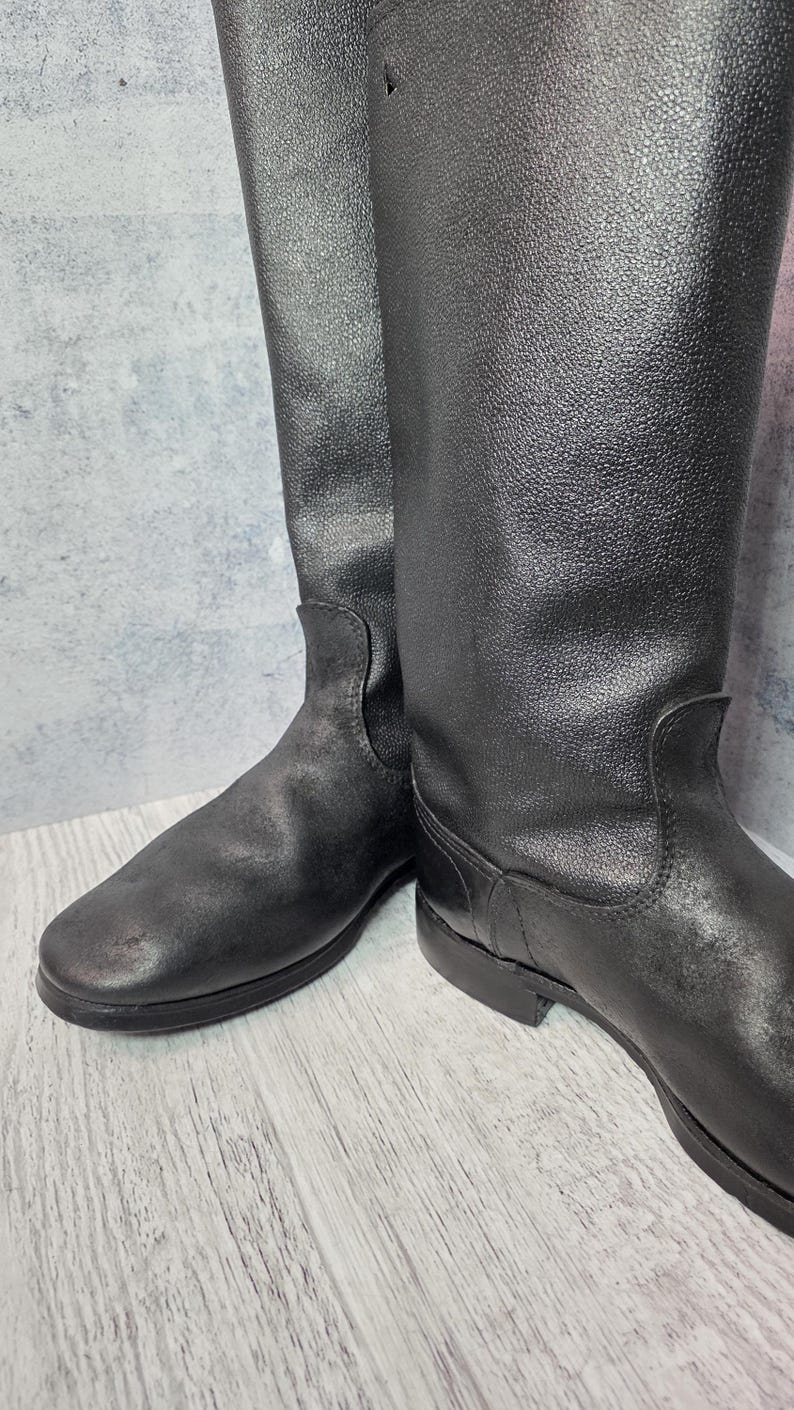 Puede incluir: Un par de botas de montar de cuero negro. Las botas tienen una ca&ntilde;a alta, una puntera redondeada y un tac&oacute;n bajo. El cuero tiene un aspecto texturizado. Las botas est&aacute;n sobre una superficie de madera clara.