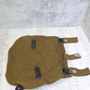 Op de afbeelding: Een bruine canvas tas met zwarte leren riemen en metalen gespen. De tas heeft een afgeronde vorm en lijkt vintage of militair te zijn. De riemen zijn vastgezet met metalen hardware.