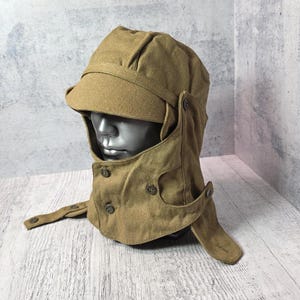 May include: A khaki-coloured military-style hat with a peak and ear flaps. The hat features a buttoned neck covering and is displayed on a mannequin head. The hat appears to be made of a robust fabric, suitable for cold weather conditions.