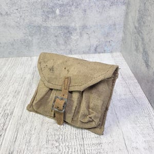 Puede incluir: Una pequeña bolsa rectangular de lona color canela con una solapa plegable asegurada por una sola correa vertical con una hebilla de metal. La tela muestra signos de desgaste y textura, con una construcción de costuras visibles y forma.