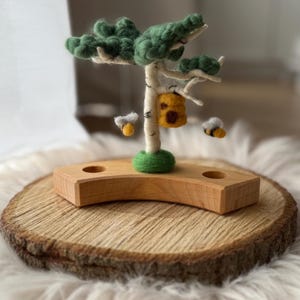 Könnte beinhalten: Eine handgefertigte Holzbaumskulptur mit einem Filz-Gründach, einem birkenähnlichen Stamm und einem Filz-Bienenstock. Zwei Filzbienen sind in der Nähe des Baumes positioniert. Die Skulptur steht auf einem Holzsockel, der auf einer Holzscheibe platziert ist.