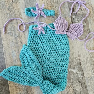 May include: A crochet mermaid costume set consisting of a teal mermaid tail with a fin, a lavender shell-shaped bikini top with tie straps, and a matching teal headband featuring a lavender starfish accent with a small white center bead.