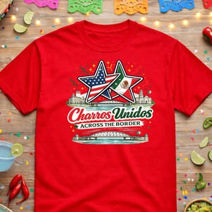 May include: Red t-shirt with a graphic featuring two stars, one with the American flag and the other with the Mexican flag. The text "Charros Unidos Across the Border" is displayed below the stars. The shirt is surrounded by festive decorations.