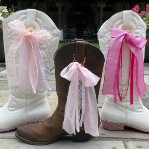 May include: Two white cowboy boots and one brown cowboy boot, each adorned with a decorative ribbon charm attached to the pull tab. The white boots feature pink rhinestone detailing along the heel and sole, while the brown boot has a lace ribbon.