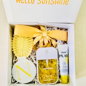 May include: A white gift box with the text HELLO SUNSHINE on the lid, containing a yellow hairbrush, a round white compact with a yellow letter N, a yellow Touchland hand sanitizer mist, and a Supergoop PLAY lip balm with a yellow satin ribbon.
