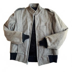 May include: A grey wool-textured bomber jacket featuring a stand-up collar, shoulder epaulettes, and a full-length front zip. The jacket includes black ribbed knit cuffs and a black ribbed hem, with a black faux fur lining visible inside.
