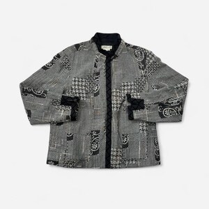 May include: A long-sleeve jacket featuring a patchwork design in shades of black, white, and gray. The garment includes a black stand-up collar, a central vertical black trim, and black quilted cuffs. The fabric displays various patterns.