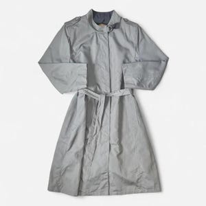 May include: A light grey trench coat featuring a high collar with a metal buckle closure, long sleeves with shoulder epaulets, and a matching tie belt at the waist. The coat has a button-down front placket and a smooth, solid-coloured fabric finish.