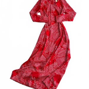 May include: A long-sleeved, floor-length red dress featuring a white geometric pattern of diagonal lines and dots. The garment has a round neckline and a fitted bodice that flows into a full-length skirt. A small brand label is visible at the inner collar.