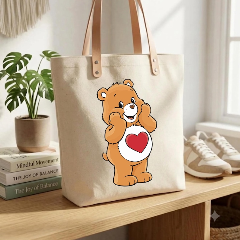 May include: A cream-colored canvas tote bag featuring a graphic of an orange bear with a red heart on its white belly. The bag has two light brown leather straps attached with silver metal rivets, providing a sturdy handle for carrying items.