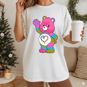 May include: A white short-sleeved t-shirt featuring a graphic of a pink cartoon bear with multicolored striped limbs. The bear has a white belly patch with a rainbow-colored heart design. The bear is waving one hand and smiling with a small heart nose.