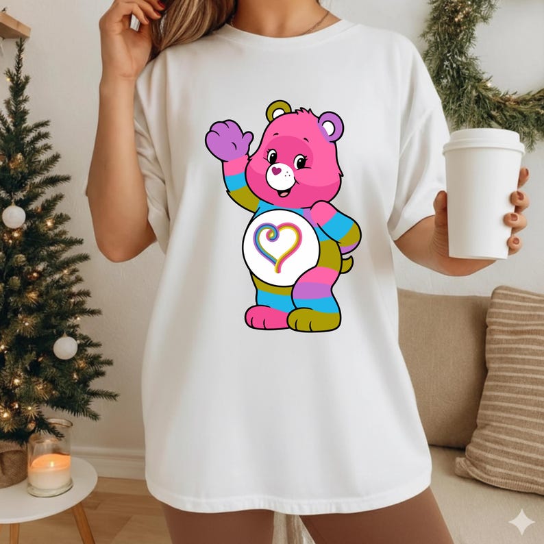 May include: A white short-sleeved t-shirt featuring a graphic of a pink cartoon bear with multicolored striped limbs. The bear has a white belly patch with a rainbow-colored heart design. The bear is waving one hand and smiling with a small heart nose.