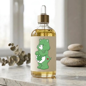 May include: A clear glass dropper bottle filled with a golden liquid, featuring a beige label with a green cartoon bear character. The bear has a white belly patch with a green four-leaf clover, a small green heart on its nose, and a red heart on its hip.