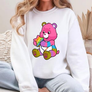 May include: A white crewneck sweatshirt featuring a graphic of a pink bear with multicolored striped limbs, holding a smiling rainbow star. The bear has a white belly patch with a rainbow heart design and purple paw pads on its feet.