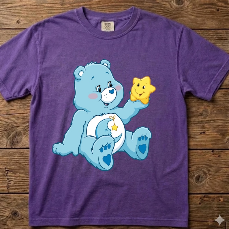 May include: A purple short-sleeved t-shirt featuring a graphic of a light blue bear with a white belly patch depicting a crescent moon and a star. The bear is holding a small, smiling yellow star in its right hand. The shirt has a crew neckline.