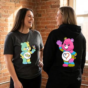 May include: A dark charcoal gray short-sleeve t-shirt featuring a pastel-colored bear with a butterfly and a black long-sleeve hooded sweatshirt featuring a bright pink bear with a rainbow heart emblem on its chest and striped limbs.