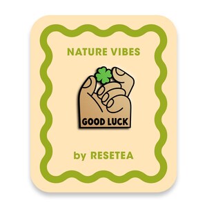 Puede incluir: Una tarjeta rectangular con un borde verde ondulado sobre un fondo crema. El texto dice NATURE VIBES en la parte superior y by RESETEA en la parte inferior. En el centro hay un pin con una mano color canela sosteniendo un trébol verde.