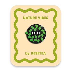 Puede incluir: Una tarjeta rectangular con un borde verde ondulado sobre un fondo crema. El texto NATURE VIBES aparece en la parte superior y by RESETEA en la inferior. En el centro hay un pin circular negro con dos ojos blancos entre hojas verdes.