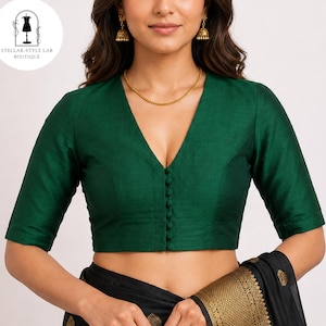 May include: A deep emerald green blouse featuring a V-neckline, elbow-length sleeves, and a front button placket with matching fabric-covered buttons. The garment is paired with a black fabric featuring gold woven patterns and gold drop earrings.