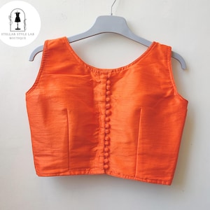 May include: A sleeveless, bright orange blouse featuring a round neckline and a vertical row of matching fabric-covered buttons down the center front. The garment has a subtle horizontal texture and is displayed on a light gray plastic hanger.