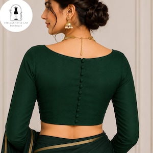 May include: A dark forest green long-sleeved blouse featuring a wide boat neckline and a vertical row of matching fabric-covered buttons running down the centre of the back. The garment is paired with a matching green fabric with a gold trim edge.