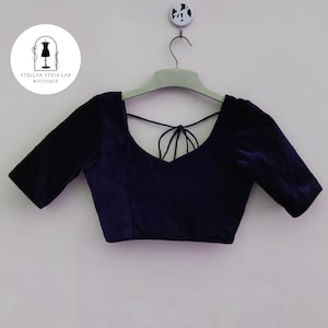 May include: A dark purple velvet blouse with elbow-length sleeves, a wide scoop neckline, and thin tie-up straps across the back. The garment is displayed on a light-colored hanger against a plain background, featuring a circular logo in the corner.
