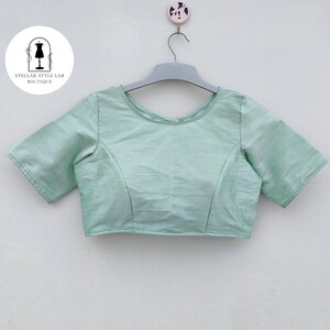 May include: A light mint green short-sleeved blouse with a round neckline and a subtle textured finish. The garment is displayed on a gray plastic hanger against a plain background, featuring a circular logo with the text STELLAR STYLE LAB BOUTIQUE.