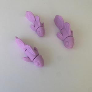 Peut inclure: Trois figurines de poissons lavande imprimées en 3D. Chaque poisson présente un design détaillé avec des nageoires et un corps segmenté. Les figurines sont disposées sur une surface blanche, mettant en valeur leur forme et leur couleur uniques.