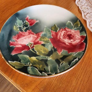 May include: A round decorative plate featuring a floral design. Two large red roses with green leaves are painted on a dark teal background. The plate has a gold rim and a white interior.