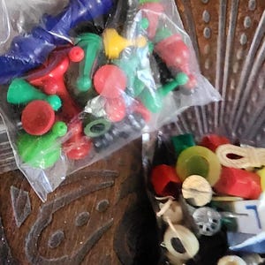 May include: Two clear plastic bags filled with colorful game pieces. The pieces are various shapes and colors, including blue, red, green, and yellow. Some pieces resemble pawns, while others are round or cylindrical.
