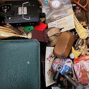 May include: A cardboard box filled with various vintage items. Includes a black metal object, a gold trophy, a green leather case, a small clock, and other decorative objects. The items are of different shapes, sizes, and colors.