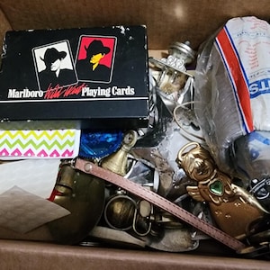May include: A box filled with various items, including a deck of "Marlboro Hotel West Playing Cards" with cowboy silhouettes, a gold-colored angel figurine, and a partially visible package labeled "FITS". Other items include a small bell and a pink strap.