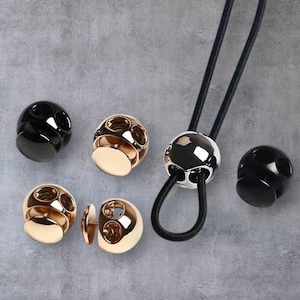 Könnte beinhalten: Eine Sammlung metallischer Kordelstopper in Gold, Silber und Schwarz. Die Stopper sind kugelförmig mit mehreren Löchern und einem flachen Boden. Eine schwarze Kordel ist durch einen der silbernen Stopper gefädelt.