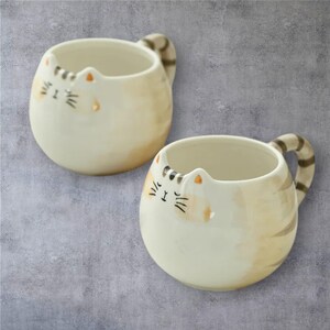 May include: Two off-white ceramic mugs with cat faces. Each mug has a rounded shape, a striped handle, and a cat face design with closed eyes, whiskers, and orange ear accents. The mugs are on a gray background.