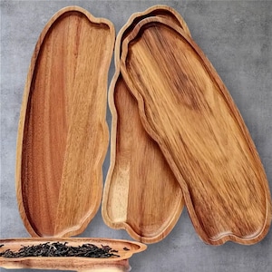 May include: Four wooden serving trays with a natural wood grain finish. The trays have an organic, wavy edge design. One tray at the bottom contains tea leaves. The trays are arranged on a grey surface.