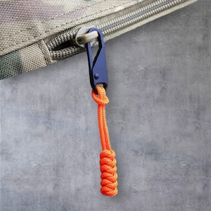 May include: A navy blue metal carabiner clip attached to a camouflage fabric zipper. An orange rope is knotted through the clip, with a decorative woven pattern at the end. The background is a textured grey surface.