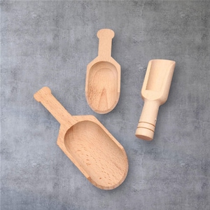 May include: Three wooden scoops of varying sizes are arranged on a gray surface. The scoops are made of light-colored wood and have a smooth finish. Each scoop has a handle and a rounded or rectangular bowl.