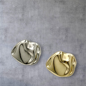 Op de afbeelding: Twee abstracte, organisch gevormde schalen. Eén is zilverkleurig, de andere goudkleurig. De schalen hebben een geribbeld, reflecterend oppervlak en staan tegen een grijze, getextureerde achtergrond. Het ontwerp is modern en minimalistisch.