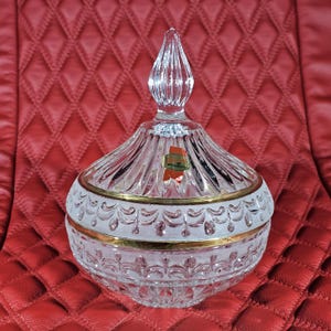 Puede incluir: Un bombonera de cristal transparente con tapa, con borde dorado y patrones decorativos grabados. La tapa tiene un remate puntiagudo. Una pequeña pegatina es visible en el lateral. El plato se encuentra sobre una superficie acolchada roja.