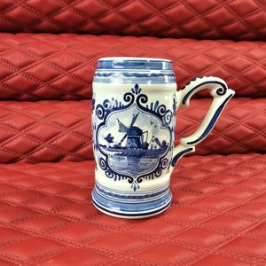Peut inclure: Une chope à bière en céramique bleue et blanche avec un motif de moulin à vent. La chope a une grande anse et est décorée de motifs floraux et de volutes. Le motif comprend un moulin à vent, de l'eau et une petite scène de village.
