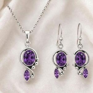 Op de afbeelding: Een zilverkleurige sieradenset met een hangerketting en bijpassende oorbellen. Elk stuk toont een grote, ovaal geslepen paarse edelsteen, geaccentueerd met kleinere edelstenen en decoratief metaalwerk. De set wordt gepresenteerd op een witte achtergrond.