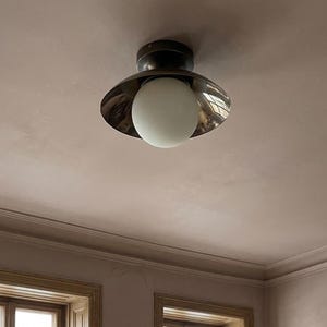 May include: A ceiling light fixture featuring a dark metal base and a reflective, flared shade. A round, white globe bulb hangs below the shade. The fixture is mounted on a light pink ceiling, with a glimpse of a window frame below.