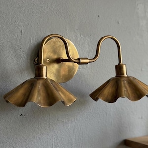 May include: A brass wall sconce with two lights. The sconce has a circular backplate and curved arms. Each light has a ruffled, bell-shaped shade. The fixture is mounted on a grey wall.