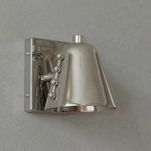 Op de afbeelding: Een gepolijste zilveren wandlamp met een klokvormige kap. De lamp heeft een rechthoekige achterplaat en een decoratieve arm met kleine bolvormige accenten. Het ontwerp is strak en modern, geschikt voor binnenverlichting.
