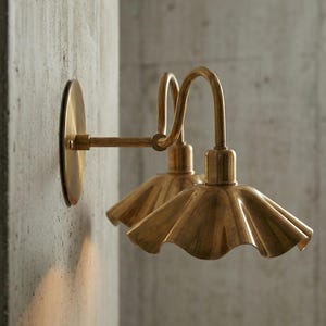 May include: Two brass wall sconces with a fluted shade design. The sconces are mounted on a concrete wall. The lamps have a curved arm and a circular base. The light fixtures have a vintage aesthetic.