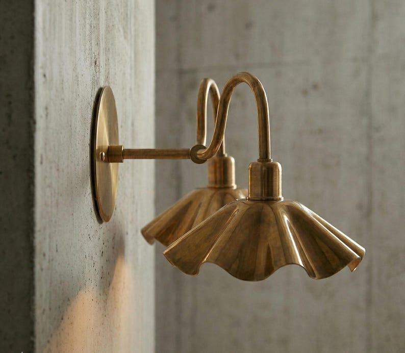 May include: Two brass wall sconces with a fluted shade design. The sconces are mounted on a concrete wall. The lamps have a curved arm and a circular base. The light fixtures have a vintage aesthetic.