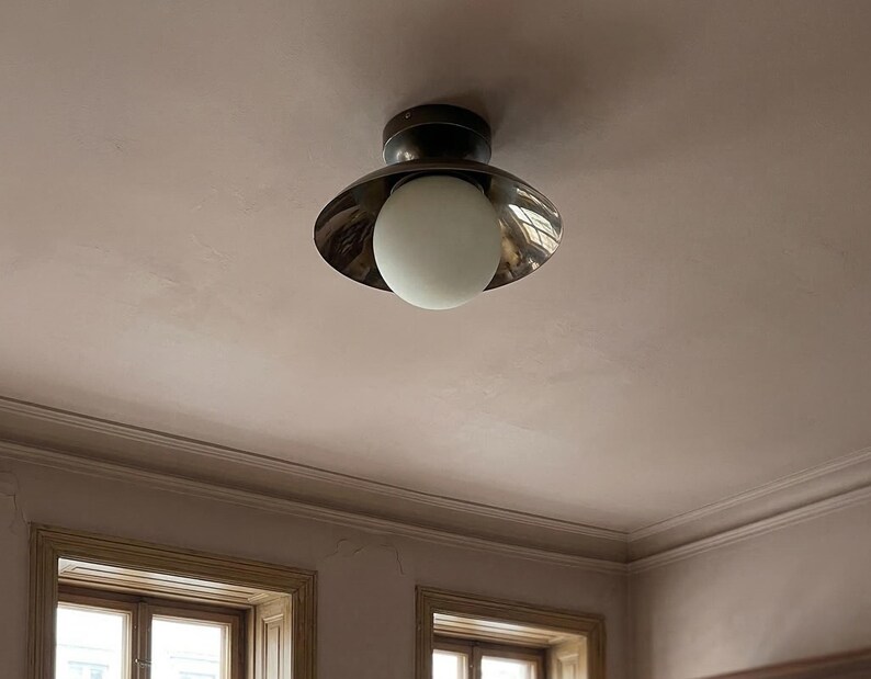 May include: A ceiling light fixture featuring a dark metal base and a reflective, flared shade. A round, white globe bulb hangs below the shade. The fixture is mounted on a light pink ceiling, with a glimpse of a window frame below.